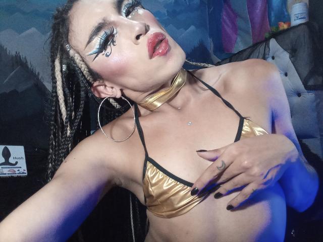 Crystalinn with braided hair, gold choker, and gold bikini top, hand on tits, red lipstick, blue eyeliner, hoop earrings, and black nail polish, in a dimly lit room.
