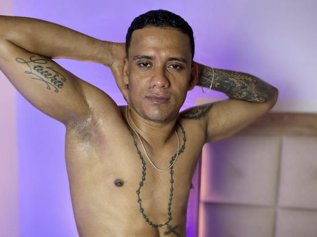 Shirtless EvaAndMarcelo with arms raised, tattoos on bicep and forearm, short black hair, brown skin, chain necklace, slight smile, purple and white background, muscular chest, small nipples.