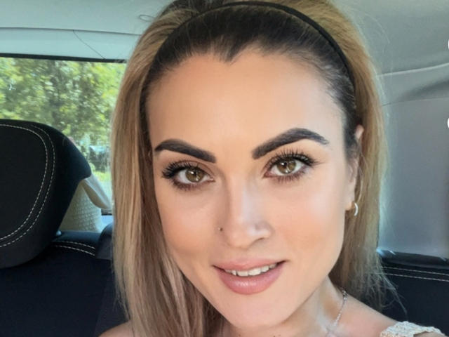 AngelDevil69Sara's face, with brown eyes, thick black eyebrows, and a slight smile, showing her bare shoulders and a headband in a car. She has small gold earrings.