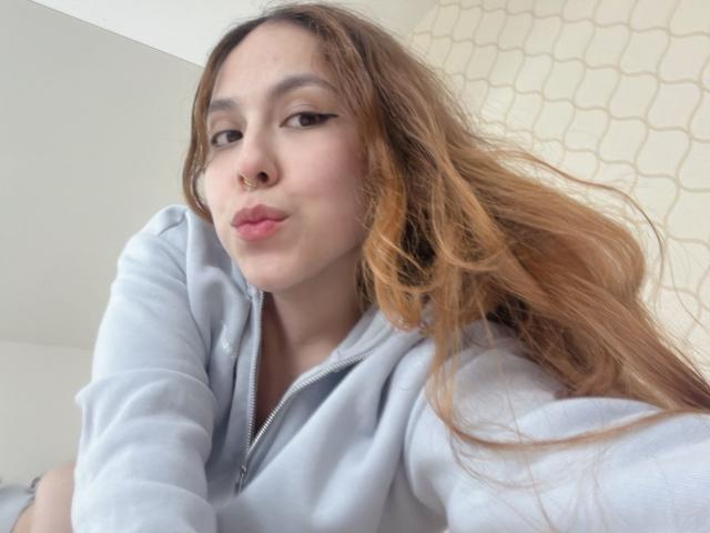 AmberlStha, con i capelli castano chiaro, labbra piene, un anello al naso e eyeliner, indossa una felpa azzurra, guarda la fotocamera con il braccio destro leggermente sollevato..