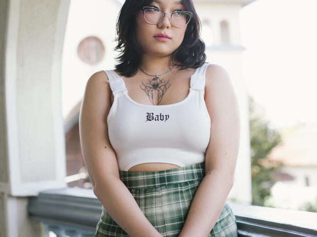 YukiLewd, met een gemiddelde borstgrootte, draagt een witte crop top met de tekst "Baby" en een groene geruite rok. Ze heeft zwart, golvend haar, een bril en een tatoeage boven haar borsten, en staat buiten..