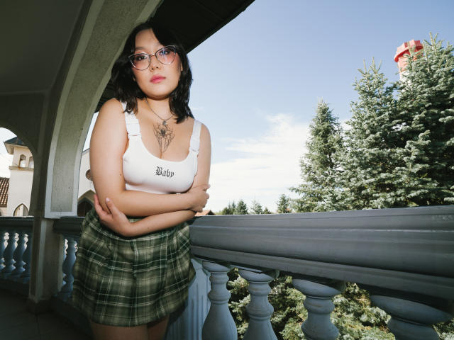YukiLewd mit Brille, weißem Tanktop, grün kariertem Rock, Armen verschränkt, stehend auf einem Balkon, grüne Bäume, klarer Himmel, weiße Brüstung, schwarzes, welliges Haar, dezenter Lippenstift, Halskette..