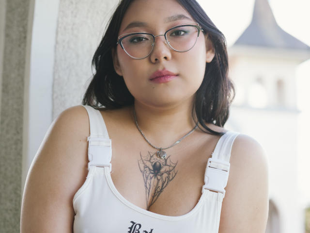 YukiLewd with glasses, brown hair, pink lipstick, white tank top, showing spider tattoo between tits, wearing necklace, standing against wall.