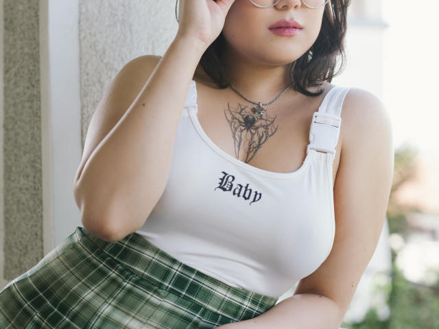 YukiLewd mit Brille, weißes Tanktop, das ihre Brüste zeigt, grüner karierter Rock, Spinnentattoo auf der Brust, Hand in der Nähe des Gesichts, weiches Licht, lässige Pose, leicht unscharfer Hintergrund..
