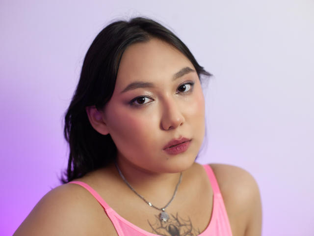 YukiLewd with pink tank top, brown eyes, and pinkish lipstick, wearing a necklace with a spider tattoo on her chest, looking slightly to the side with a neutral expression.