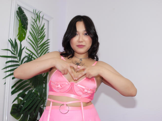 YukiLewd with black hair, pink lingerie, and heart hands, showing tattoos, pink nipples, and pink pussy outline, standing by green plant.