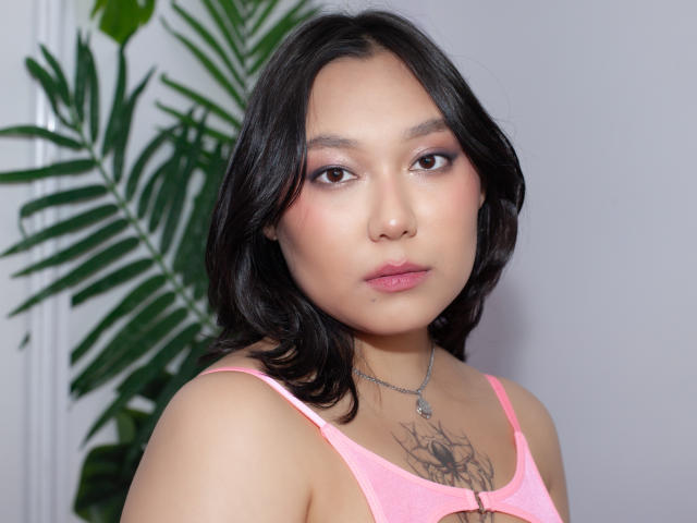 YukiLewd mit mittelbrauner Haut, schwarzen, welligen Haaren, einem pinken Tanktop, einem Tattoo auf der Brust, einer Halskette und dezentem Make-up, blickt leicht nach rechts mit einem neutralen Gesichtsausdruck..