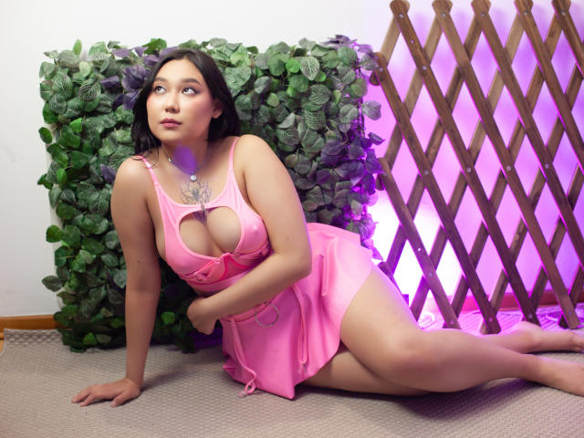 YukiLewd avec de grandes seins, vêtue d'une robe rose, assise sur un tapis, avec un mur de feuilles vertes en arrière-plan, une structure en bois avec un éclairage violet, regardant vers le haut, une main sur le sol, de longs cheveux noirs..