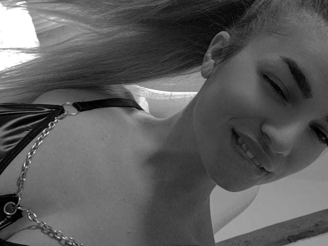 Black-and-white selfie of IngridI with long hair, smiling, wearing a leather harness with a chain, showing her shoulders and upper chest, her tits partially visible.