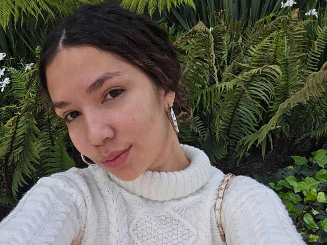 KendallPiierce69, with puffy lips and brown eyes, wearing a white cable knit sweater, has curly brown hair and small hoop earrings, standing in front of green ferns.