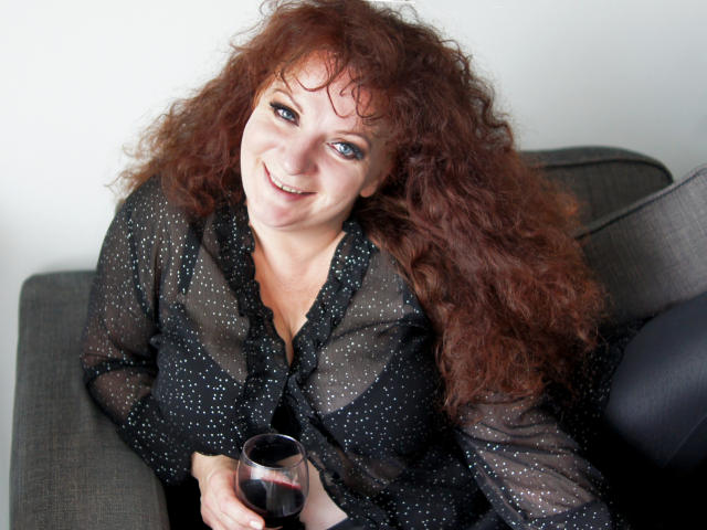 AliceXPerfect with curly red hair, black polka dot blouse, holding a glass of red wine, smiling at the camera, sitting on a gray couch.