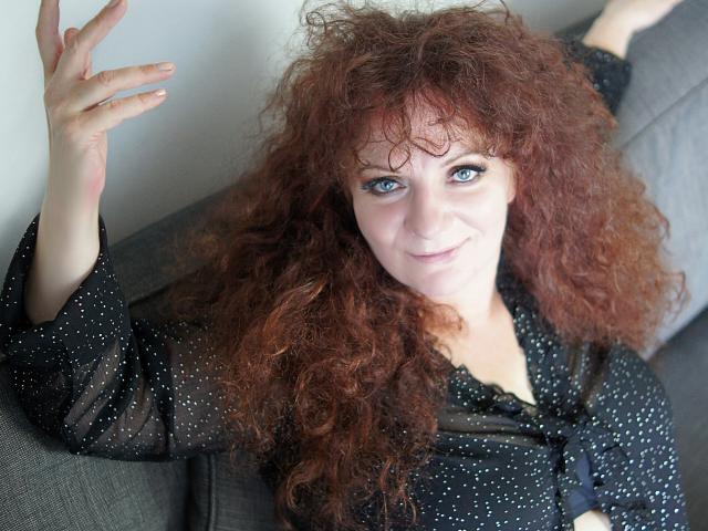AliceXPerfect with curly red hair, blue eyes, and a slight smile, wearing a black polka dot shirt, raising one hand. Her tits are small and she's on a gray couch.