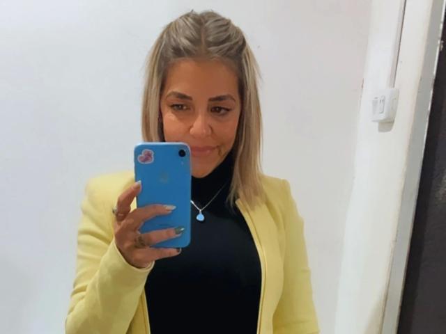 GuadaFeliz-hot with blonde braids, yellow cardigan, black turtleneck, holding blue phone, smiling at mirror, showing ring on finger, necklace with small pendant, looking cute.