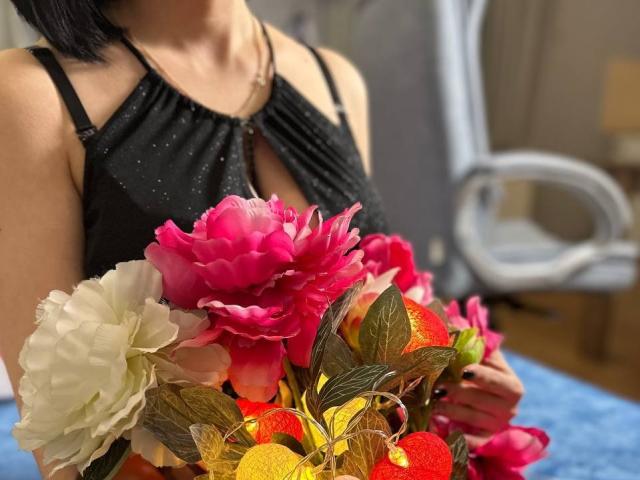 Janel's black dress with a keyhole, holding a colorful bouquet of pink, white, and orange flowers. Her black bob hair is partially visible. Her ass is slightly exposed.