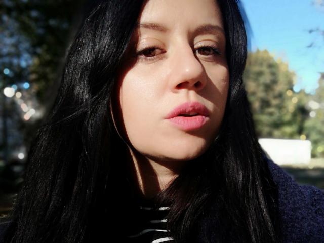 SamanthaApple's face with long black hair, pink lips, and subtle makeup, wearing a striped shirt and dark jacket, looking slightly to the side in bright sunlight.