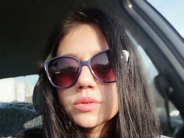 SamanthaApple with dark straight hair, wearing purple sunglasses, has pinkish lips, and a light skin tone, sitting in a car with a green headrest behind her.