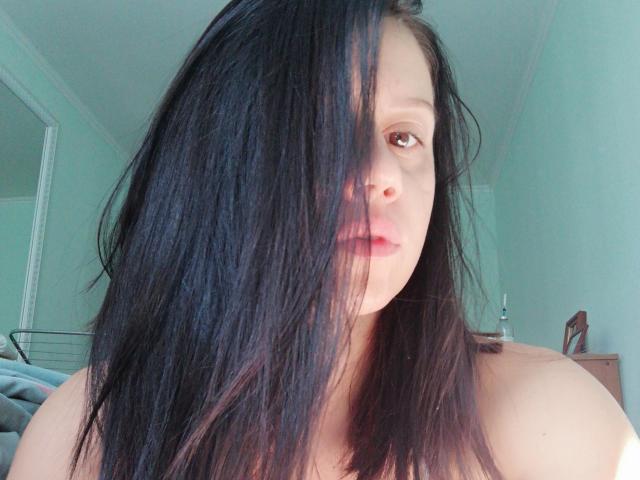 SamanthaApple with long black hair covering one eye, light skin, and pink lips, looking slightly to the side, bare shoulders visible, in a sunlit room.