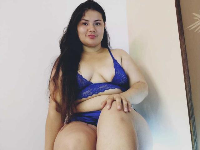 ZoeyClay, plus-sized with long black hair, wearing blue lace bra and matching panties, sits with one knee up, looking at camera, slightly smiling, showing her tits and ass.