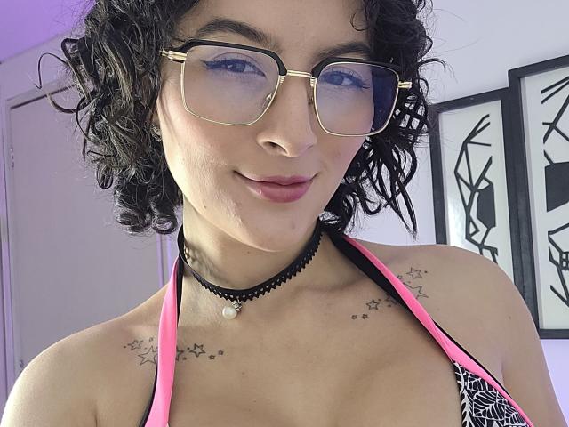 GabrielaMoore with curly black hair, gold-framed glasses, pink and black halter top, small star tattoos on shoulders, black choker with pearl, smiling at camera.