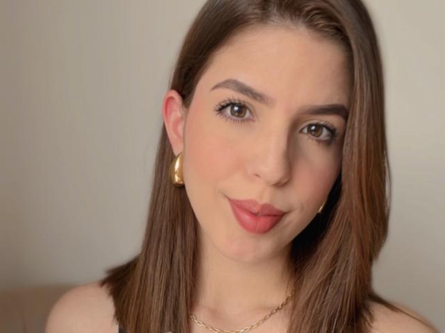 NatyBakers tiene el cabello castaño, aretes de aro dorados y una sonrisa sutil con lápiz labial rojo. Se pueden ver sus ojos marrones y su piel clara. Lleva un collar dorado..