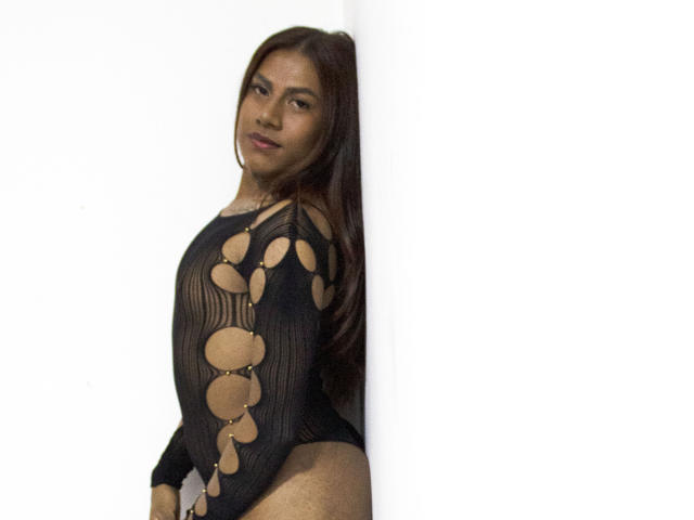 KimmySolange with brown hair, wearing a black mesh top with circular cutouts, showing her tits and ass, standing against a white wall, smiling subtly.