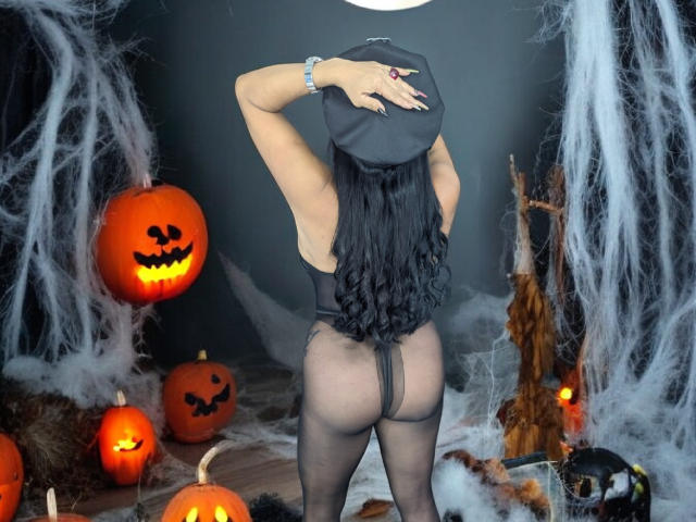 ScarletteStones, com longos cabelos pretos, uma tanga preta e meias-calças pretas transparentes, cobrindo a cabeça com um chapéu preto, está em frente a decorações de Halloween, incluindo abóboras esculpidas..