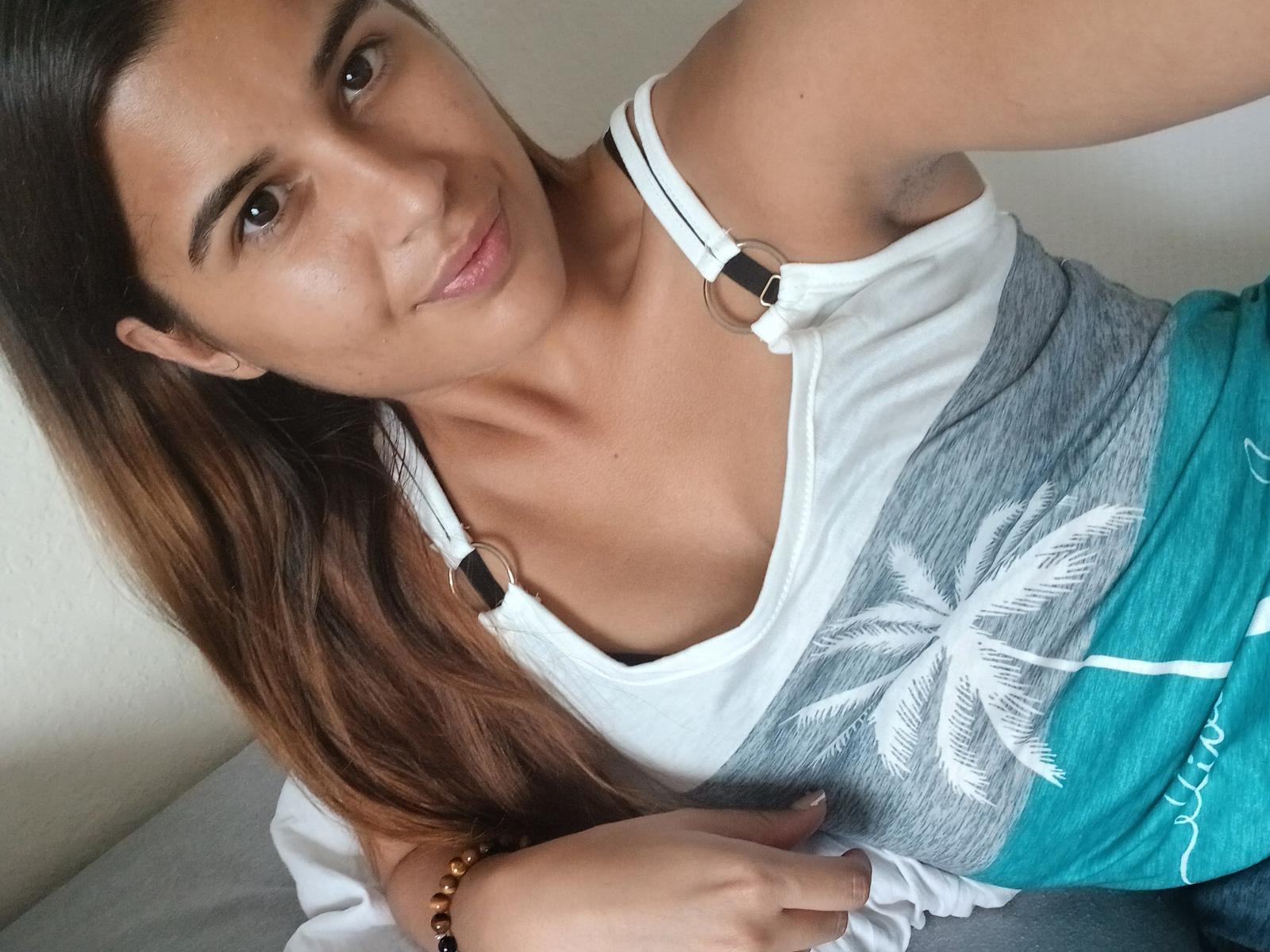 Rosadi with long brown hair, tan skin, and brown eyes, wearing a white tank top with gray palm print and turquoise shorts, smiling at the camera, showing small tits.