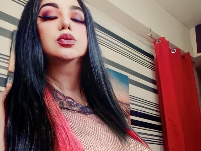 MistressQueenDominaTS with long black hair, pink eyelids, red lipstick, and mesh top, pouting with visible boob outline, pink nipple showing through, red curtains in background.