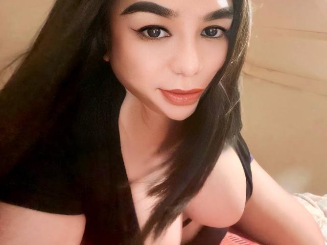 MistressQueenDominaTS with dark hair, thick eyebrows, and full lips, wearing a black top, showing her big boobs in a selfie. She has a cute, sexy look.