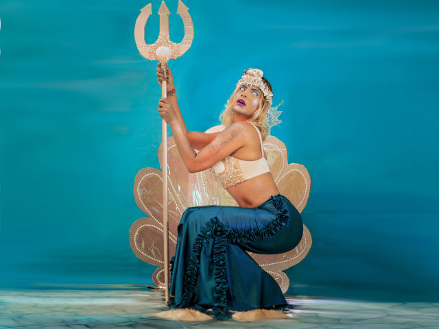 ChantalSex69 with blonde hair, blue eyes, and tattoos, squats holding a trident, wearing a white crop top, blue ruffled skirt, and flower crown, showing her ass.
