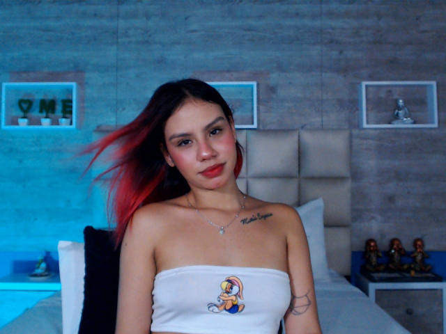 SussyGray with red and black hair, wearing a white tube top with a cartoon character, has small tits, a tattoo on her right arm, and a necklace.