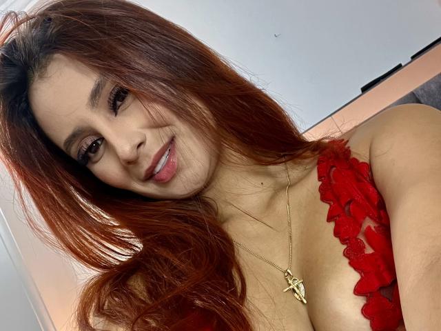MyaSpecter has long red hair, a cute smile, and is wearing a red lace top, showing her small tits and a gold necklace with an 'A' pendant.