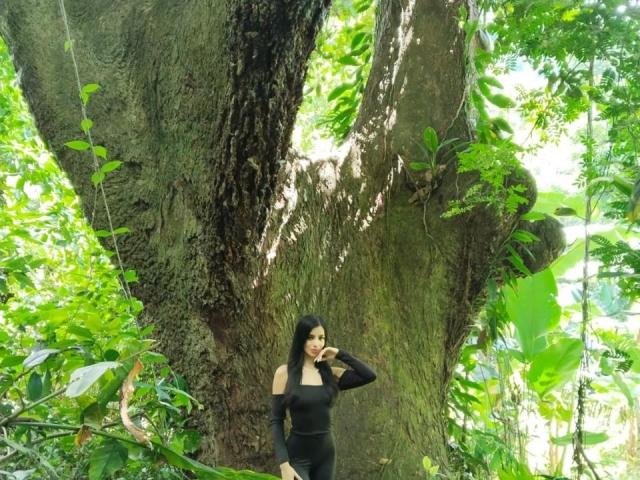 ShantalTurner with black off-shoulder top and black pants, standing in front of huge tree, touching her mouth, green jungle foliage around, long black hair, looking curious.