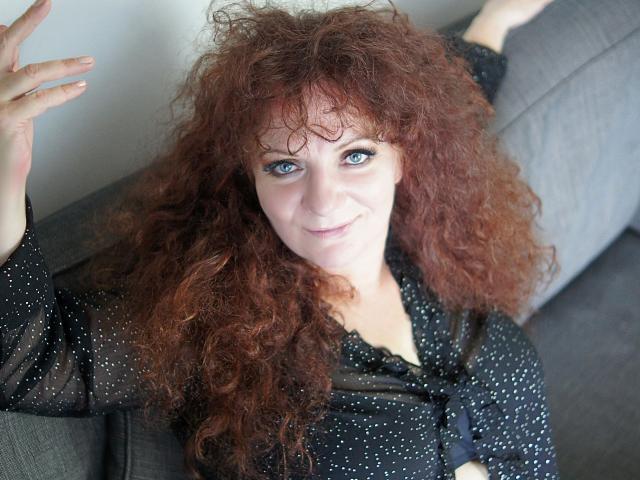 Delight-Alice with curly red hair, blue eyes, and a slight smile, wearing a black polka-dot top, sits on a gray couch, raising one hand.