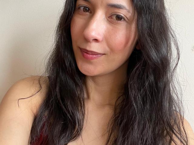 AnneStone has long, wavy black hair, a light brown skin tone, and a subtle smile with pink lips. She's wearing a red top, showing her small, round tits.