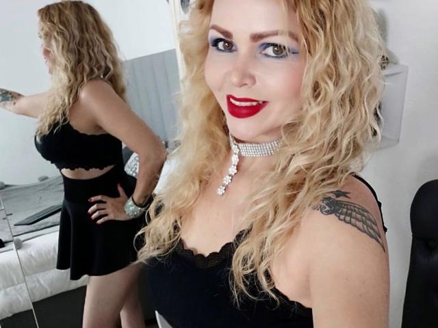 AnnaRichie with wavy blonde hair, red lipstick, black dress, and choker, showing her back and tattooed arm in mirror selfie, hands on hips, smiling confidently.