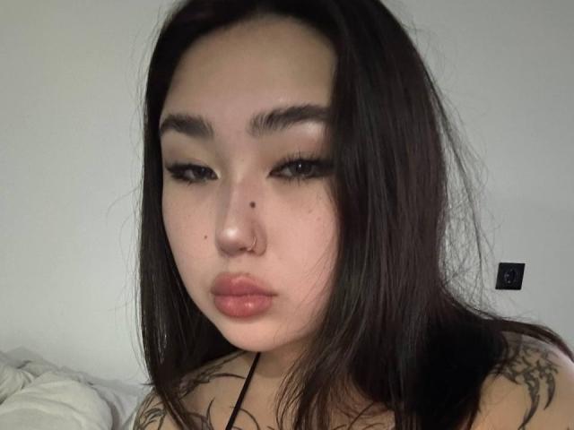 YurikoMay has long dark hair, thick eyebrows, long eyelashes, a nose ring, and full pink lips. Her tattoos are visible on her shoulders. She looks down slightly.