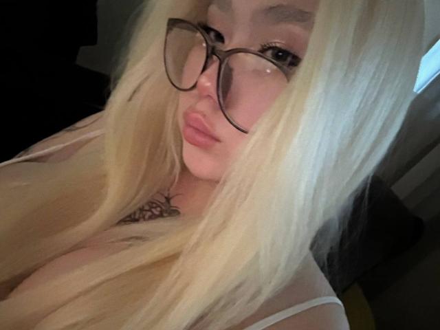 Blonde YurikoMay with big tits, wearing glasses, pouty lips, and a tattoo under her neck, looking at the camera. Her long hair covers part of her face.