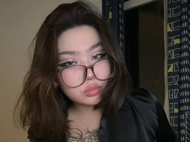 YurikoMay with puffy lips, dark wavy hair, and big glasses, wearing a black satin shirt, showing a tattoo on her chest. Her eyes are half-open.