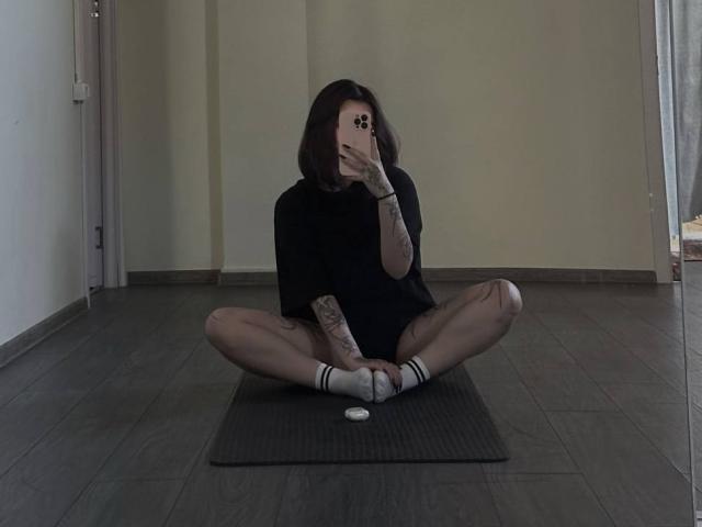 YurikoMay, sitting on a mat, legs spread, black shirt, white socks, holding phone, tattoos on arm, ass exposed, taking a selfie, dark room, wooden floor.
