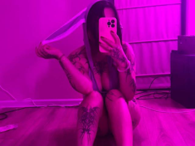 YurikoMay with tattoos, pink-lit room, holding white fabric over head, sitting on floor, taking selfie with iPhone, showing her tits, tattoos on arms and legs, wooden floor.