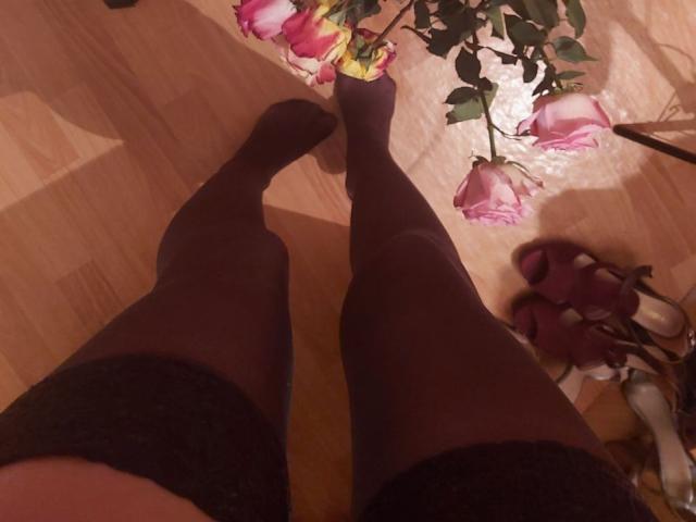 ElzaKomx's legs in black lace-top stockings, standing on a wooden floor, with pink roses and green leaves overhead, and pink high heels to the right.