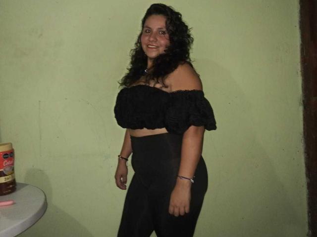 Ana Jimenez, con i capelli ricci e scuri, sorride, indossa una maglietta nera con una spalla scoperta e pantaloni neri a vita alta, ed è in piedi vicino a un piccolo tavolo con un barattolo di burro di arachidi..