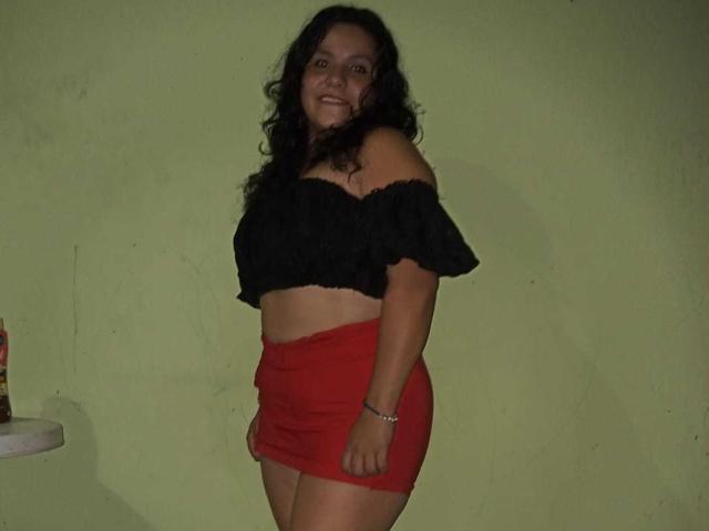 Ana Jiménez, con cabello oscuro y rizado, sonriendo, vestida con una blusa negra con un hombro descubierto y una falda roja mini, mostrando su trasero y un ligero bronceado, está de pie en una habitación con poca luz..