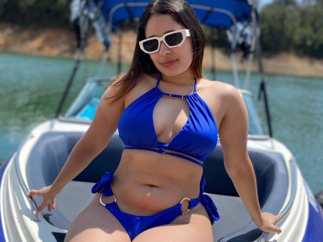 ElinaLeducc in a blue bikini, showing big tits and a soft belly, sitting on a boat with white sunglasses, water in the background, and a blue boat cover.