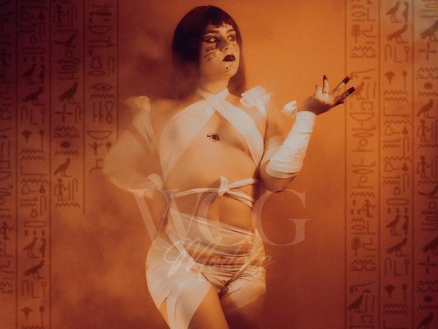 IsabelaCooper with dark bob wearing white, crisscrossed wrap-around, exposing tits, belly button, and pussy, against an orange, ancient Egyptian-themed backdrop, with arm raised, eyes closed.