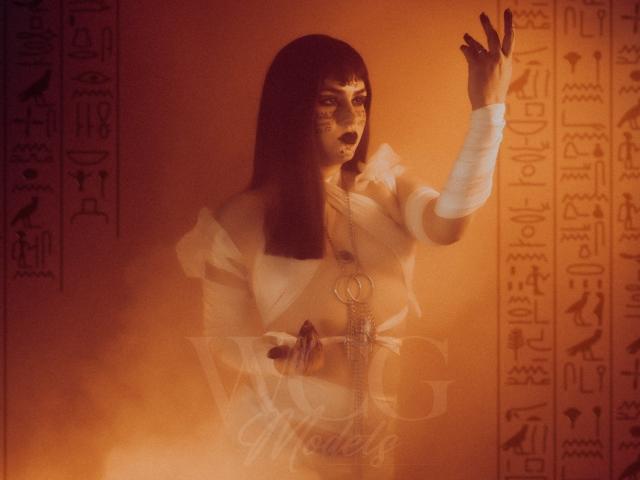IsabelaCooper with dark bob, Egyptian-style makeup, white dress, bandaged arm, holding an ankh, raising hand, orange hazy background, ancient Egyptian vibe, serious expression.
