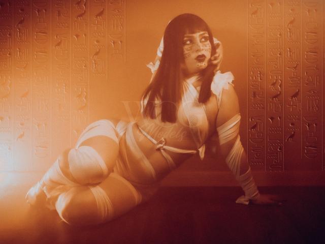 IsabelaCooper is on all fours, wrapped in white bandages, with Egyptian-style face paint and dark hair, in a warm, orange-toned, ancient Egyptian-themed photo.