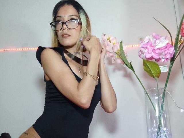 Veroo69 with blonde hair, glasses, black tank top, pulling strap, showing small tits, necklace, tattoo, and pink flowers in glass vase. Red LED strip light in background.