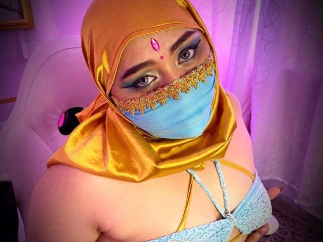 AishaGhulam con un hijab de satén amarillo, una máscara de encaje azul y un sujetador a juego, mostrando sus pechos, con una gema rosa en la frente, mirando hacia arriba con un maquillaje de ojos azul llamativo..